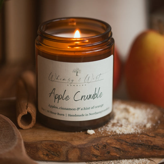 A lit amber glass jar candle labeled 'Whimsy & Wist Candles' in the scent 'Apple Crumble,' styled on a wooden surface with flour, a cinnamon stick, and a blurred apple in the background.