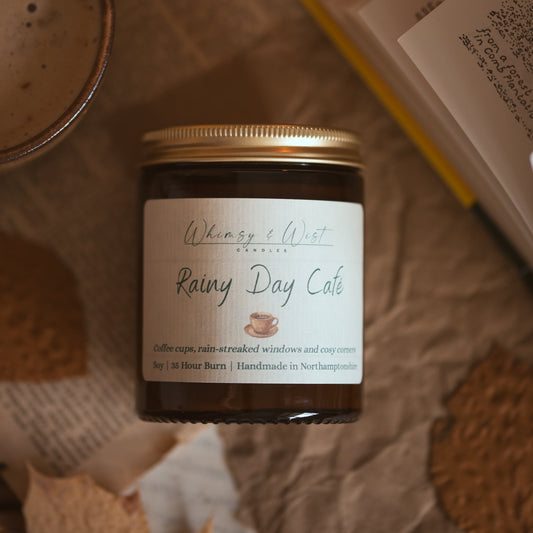 A glass jar candle with a gold lid and a white label reading 'Whimsy & Wist Candles' and 'Rainy Day Cafe is styled on crumpled paper with an open book and a coffee cup nearby.