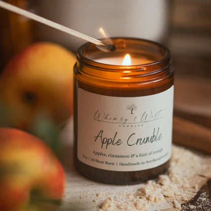 An amber glass jar candle labeled 'Whimsy & Wist Candles Apple Crumble' lit with a wooden match, set on a rustic wooden surface with apple slices and crumbs in the background.