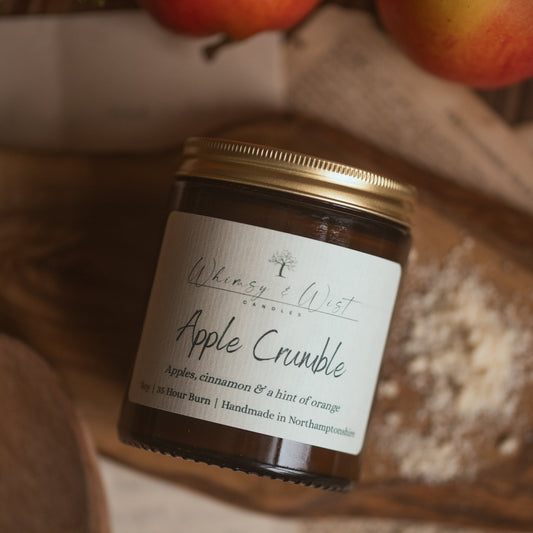 A jar candle labeled 'Apple Crumble' by Whimsy & Wist Candles, styled on a rustic wooden surface with whole apples in the background.