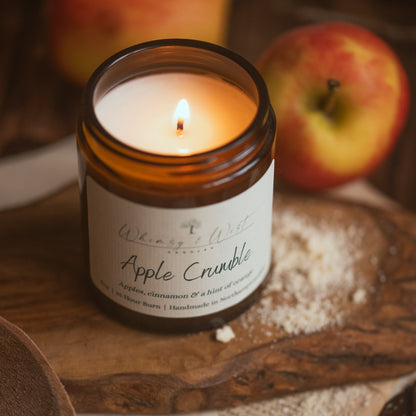 A lit amber glass jar candle labeled 'Whimsy & Wist Candles, Apple Crumble' sits on a wooden board with flour, cinnamon sticks, and blurred apples in the background.