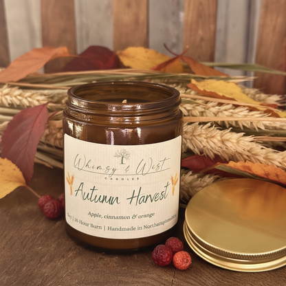 Candle jar labeled 'Autumn Harvest' with autumn themed decor on a wooden surface