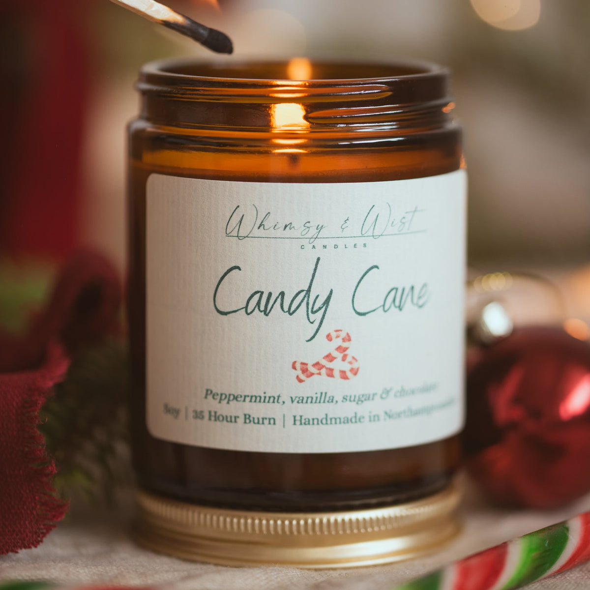 Candy Cane soy candle being lit with a match, standing on its gold lid beside a red bauble