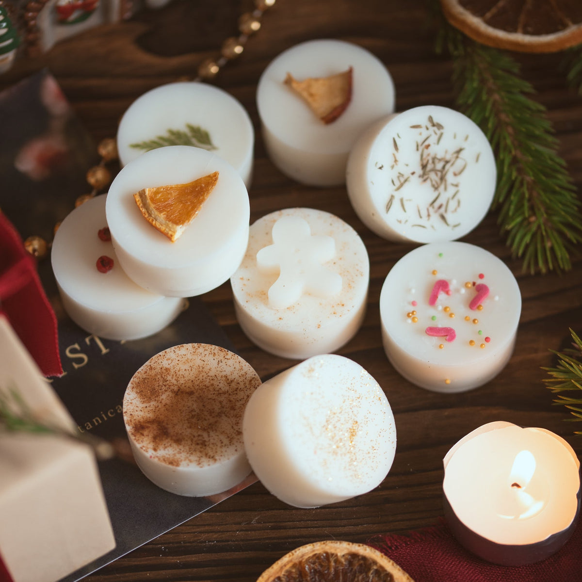Nine 'Scents of Christmas' wax melts topped with dried orange, gingerbread men, candy canes, pine and spices, beside a lit tea light