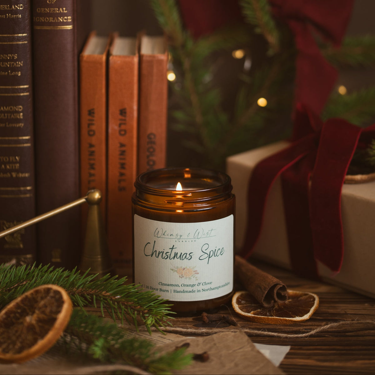 Lit Christmas Spice soy candle beside vintage books, a brass snuffer, pine and a gift tied with red velvet ribbon