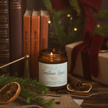 Lit Christmas Spice soy candle beside vintage books, a brass snuffer, pine and a gift tied with red velvet ribbon