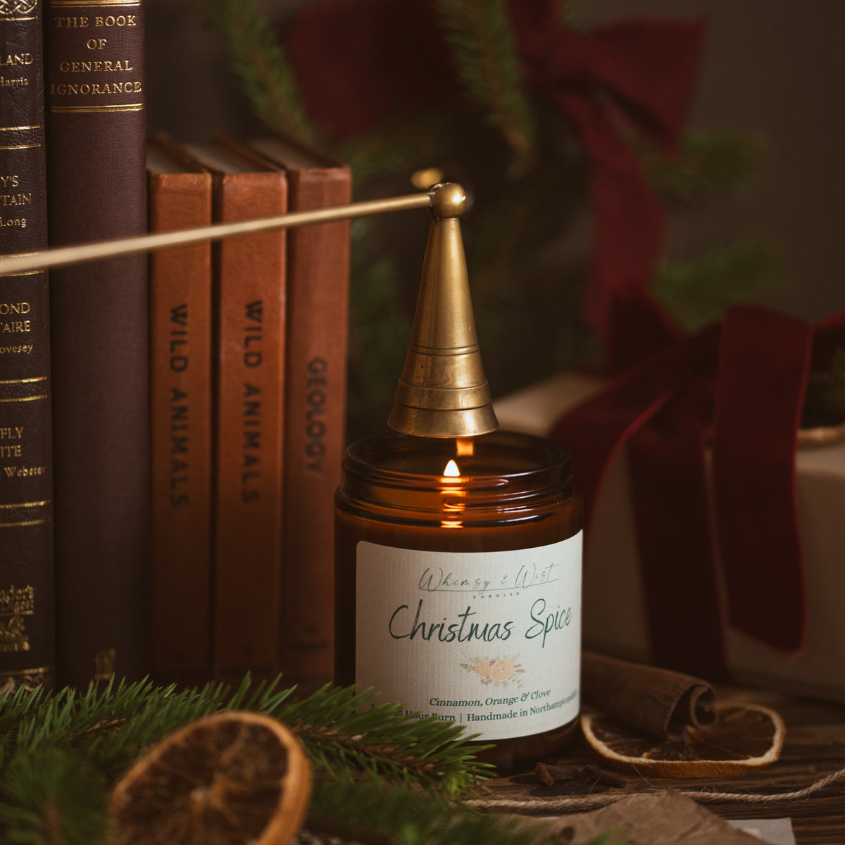 Christmas Spice soy candle being put out with a brass snuffer in front of vintage books and red velvet ribbon