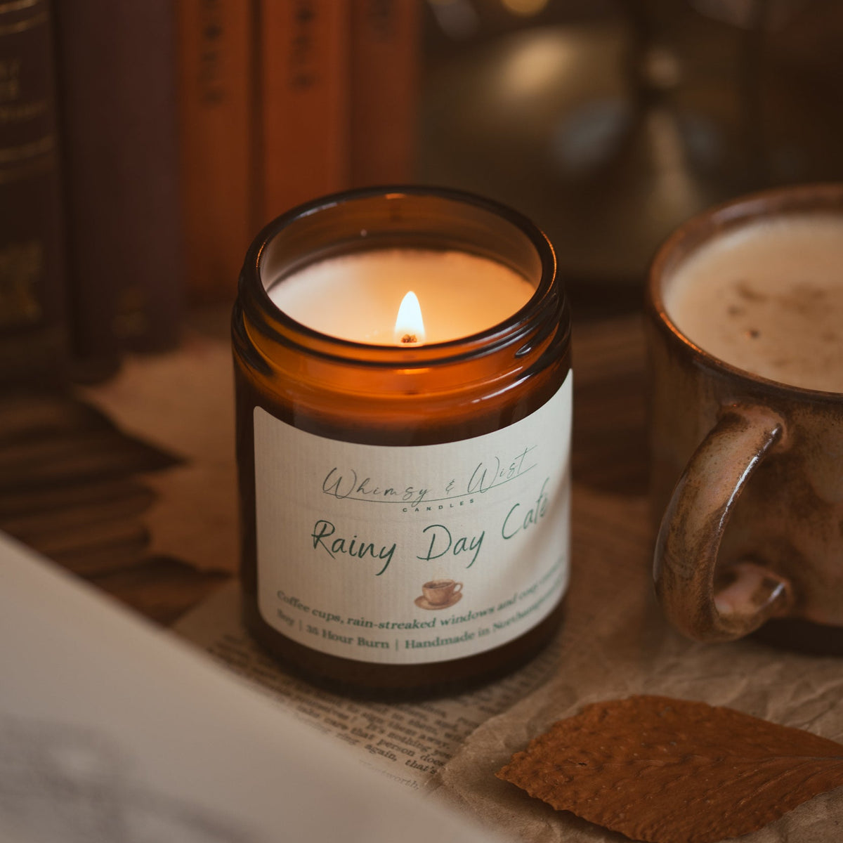 An amber glass jar candle labeled 'Whimsy & Wist Rainy Day Cafe' lit and set on a tabletop next to a mug, with books and a candle in the background.