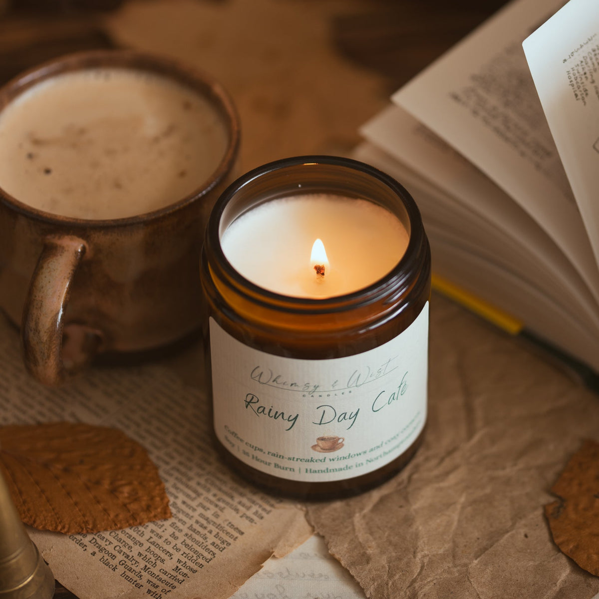 A lit amber glass jar candle labeled 'Rainy Day Cafe' sits on a table with an open book, a mug of coffee, and scattered book pages in a cozy, warm setting.