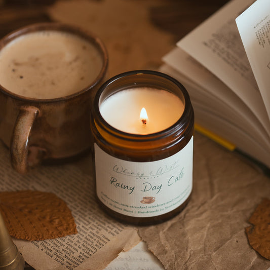 A lit amber glass jar candle labeled 'Rainy Day Cafe' sits on a table with an open book, a mug of coffee, and scattered book pages in a cozy, warm setting.