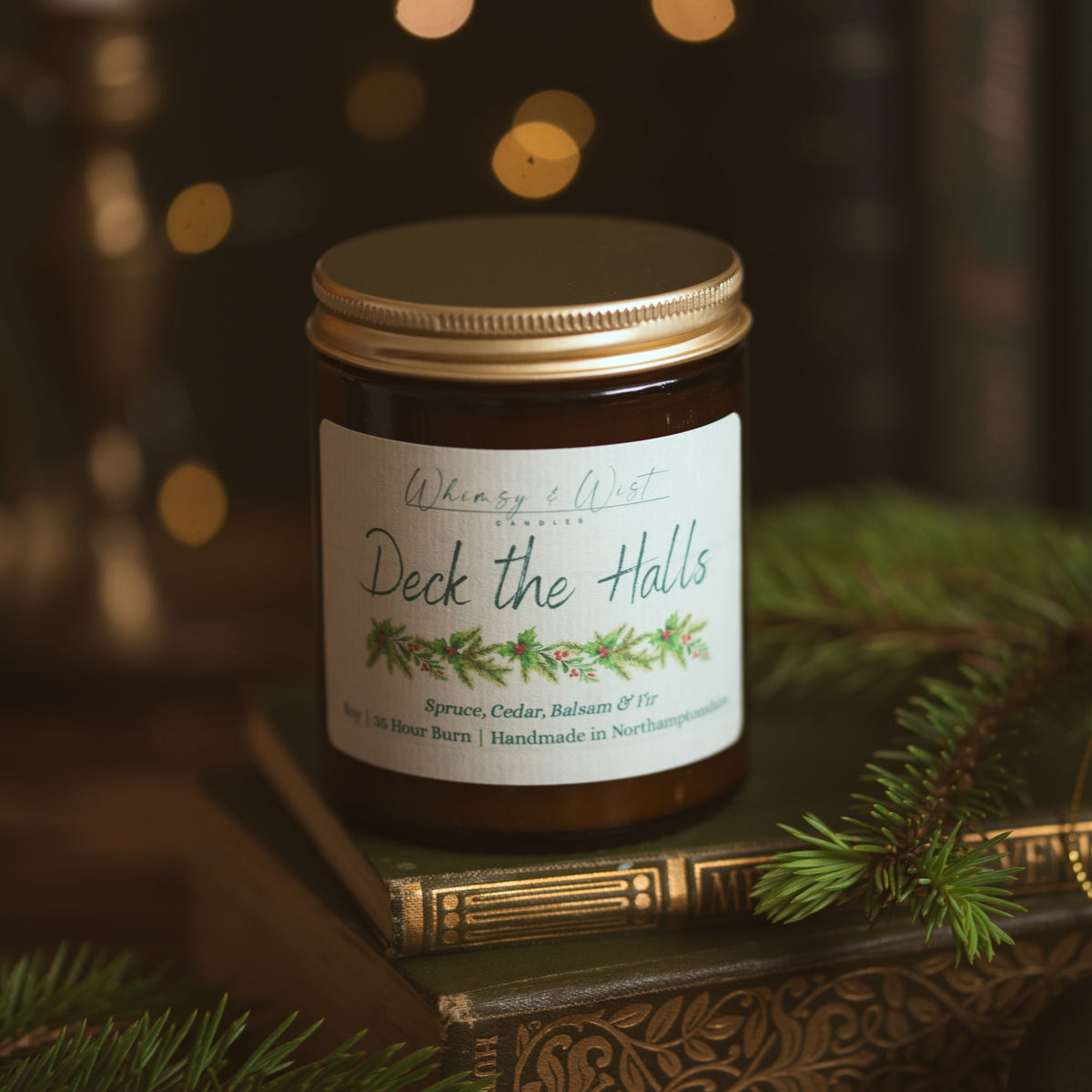 Whimsy & Wist Deck the Halls soy candle with gold lid on vintage green books, surrounded by fresh pine