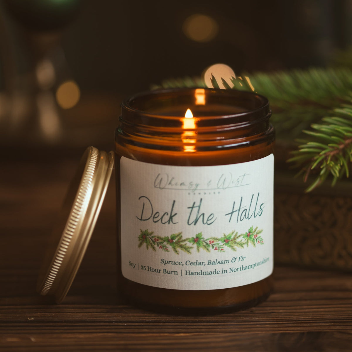 Lit Deck the Halls soy candle on a wooden table with its gold lid propped beside it and pine branches