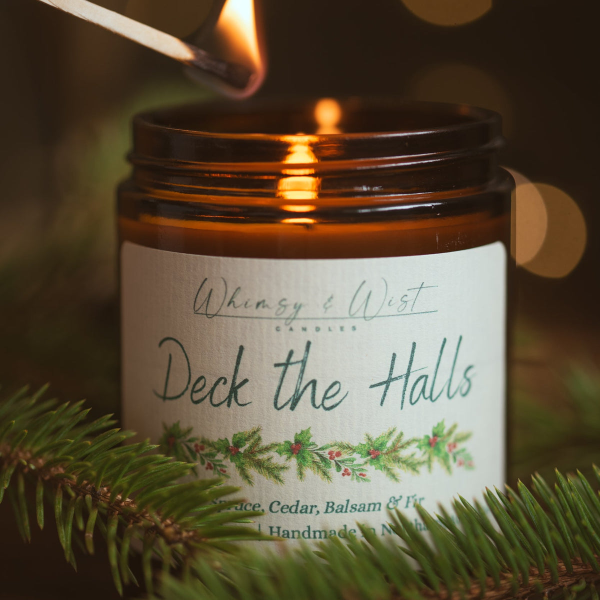Deck the Halls soy candle being lit with a match, framed by fresh pine needles