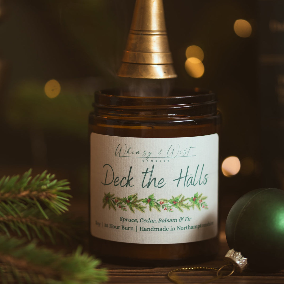 Deck the Halls soy candle being put out with a brass snuffer, next to a green Christmas bauble