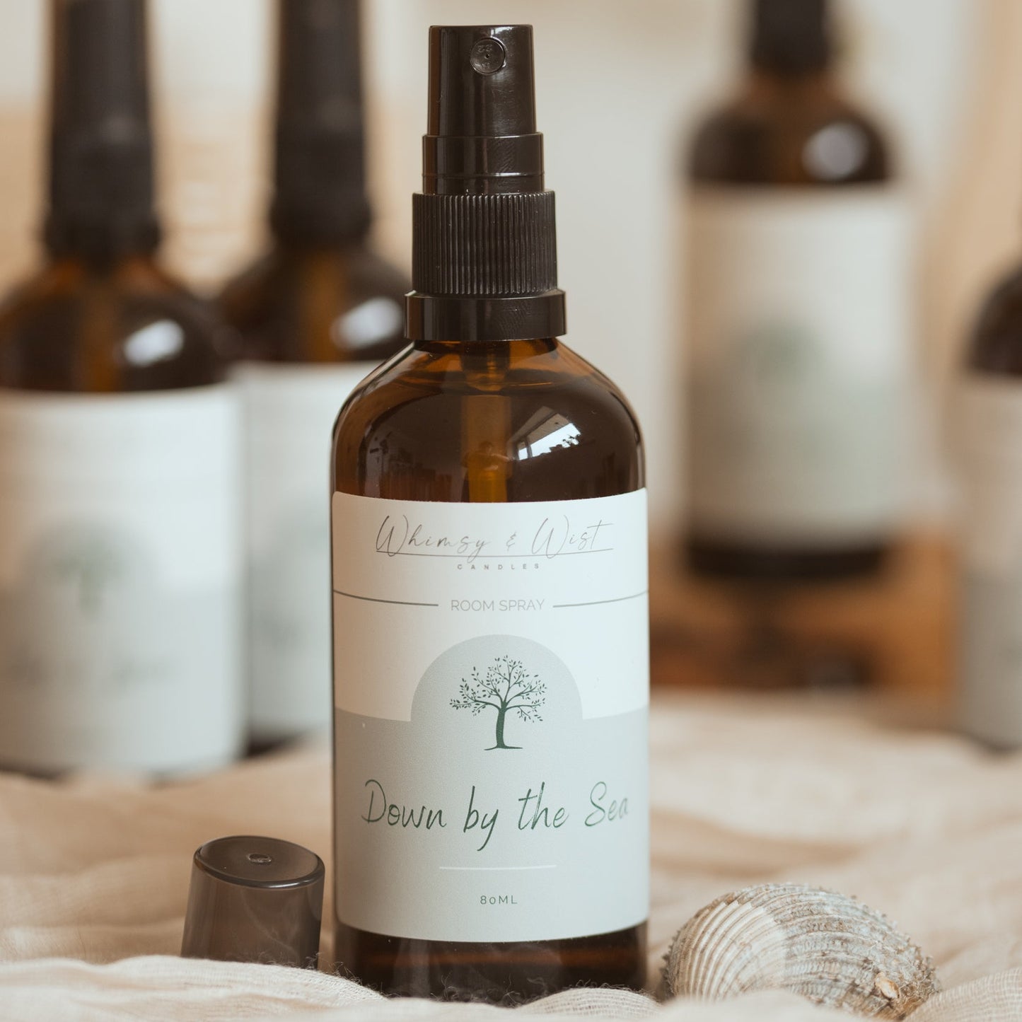 An amber glass spray bottle with a black spray top and a white label reading 'Whimsy & Wist' and 'Down by the Sea', styled on a soft beige fabric with a cap nearby and other blurred bottles in the background.