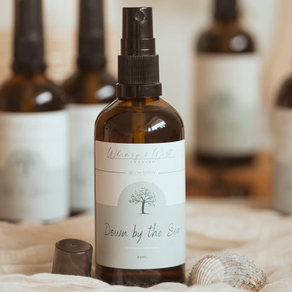 An amber glass spray bottle with a black spray top and a white label reading 'Whimsy & Wist' and 'Down by the Sea', styled on a soft beige fabric with a cap nearby and other blurred bottles in the background.