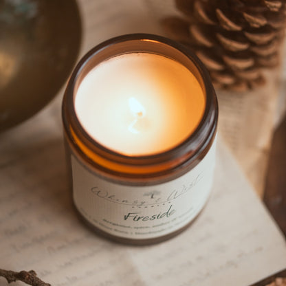 Candle in a jar labeled 'Fireside' on a surface with pine cone and branch