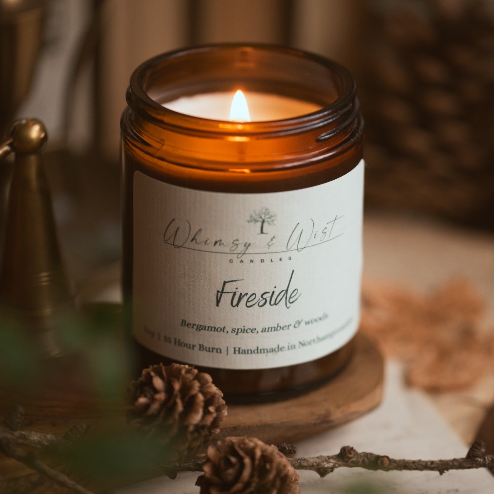Candle labeled 'Fireside' from 'Whimsy & Wist' on a wooden surface with pinecones and branches.