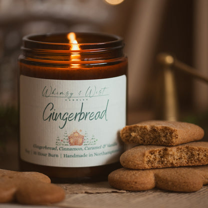 Lit Gingerbread soy candle in an amber jar beside a stack of broken gingerbread biscuits