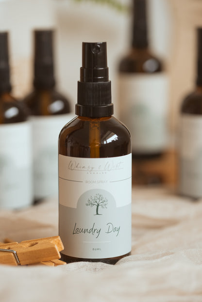 An amber glass spray bottle labeled 'Laundry Day' by Whimsy & Wist set on a beige fabric surface with wooden clothespins and blurred matching bottles in the background.