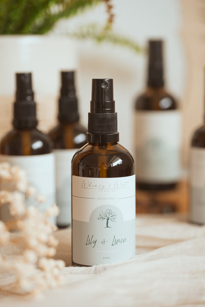 An amber glass spray bottle with a black pump and a white label reading 'Lily + Linen' stands in the foreground on a soft fabric surface, with several matching bottles blurred in the background.