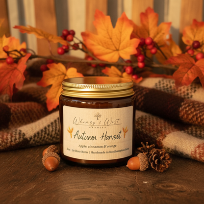 Candle jar labeled 'Autumn Harvest' surrounded by autumn decorations on a wooden surface.