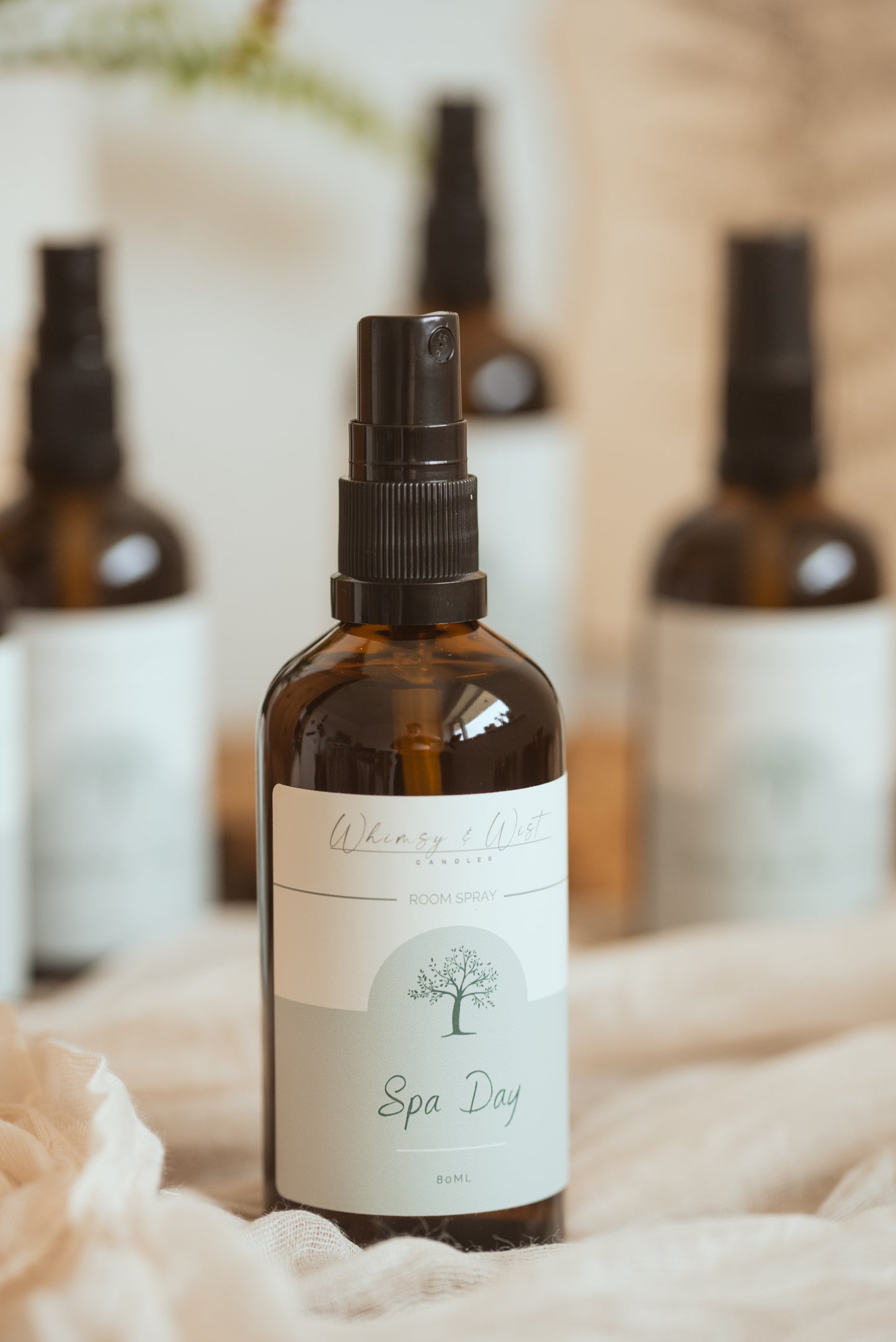 An amber glass spray bottle labeled 'Whimsy & Wist' and 'Spa Day' in the foreground, with three similar bottles blurred in the background on a soft beige fabric surface.