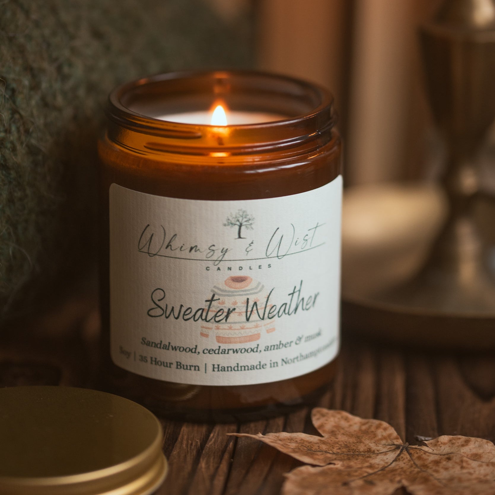 Candle labeled 'Sweater Weather' with a warm glow, surrounded by autumn leaves and a blurred candle in the background.