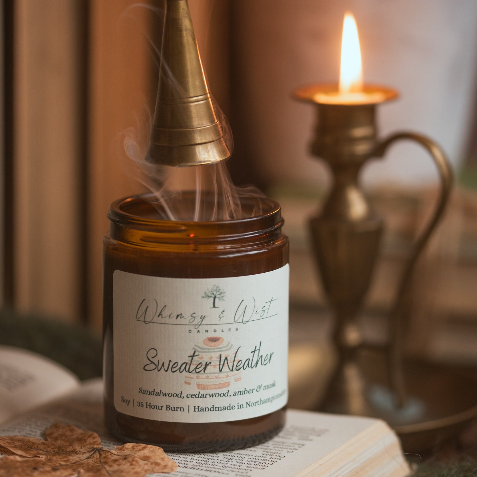 A 'Sweater Weather' candle with a brass snuffer on an open book, surrounded by a cosy setting.