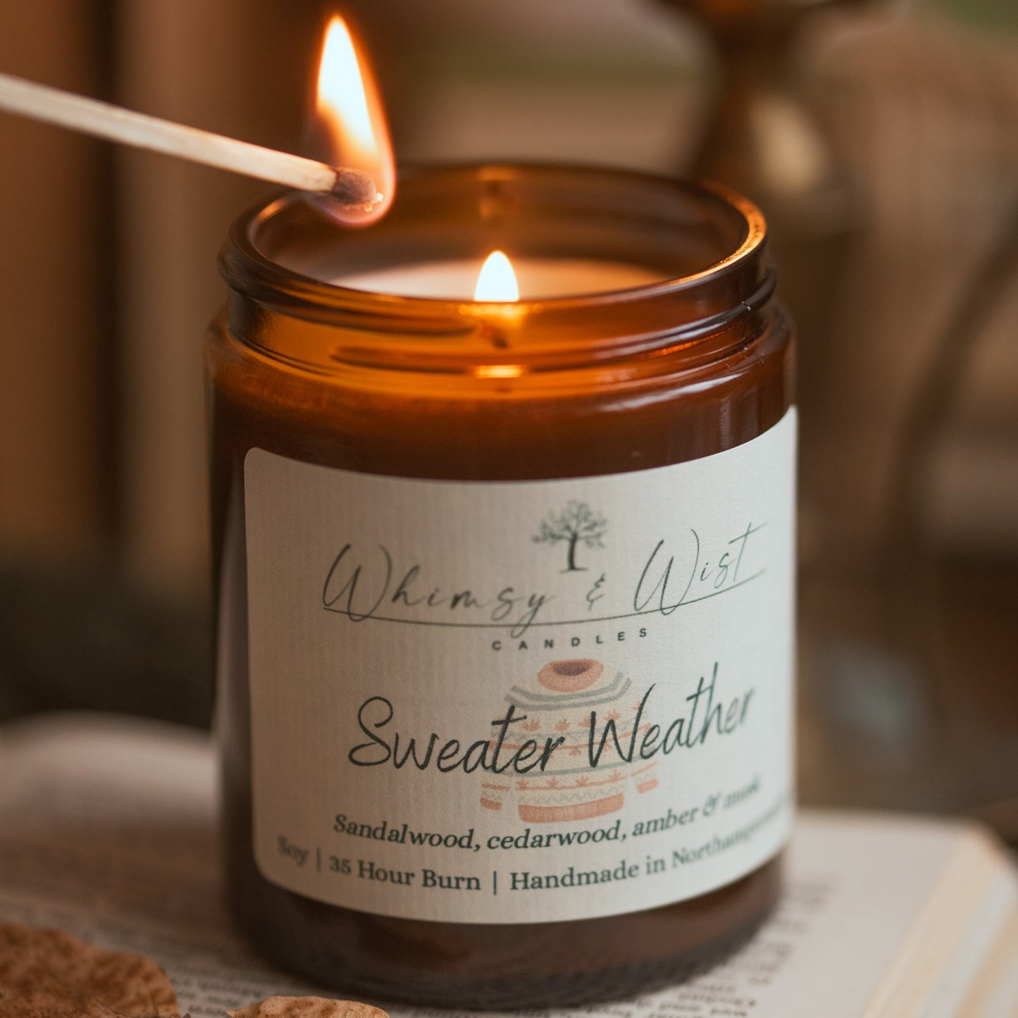 Candle labeled 'Sweater Weather' from 'Whimsy & Wist' on an open book with a leaf.