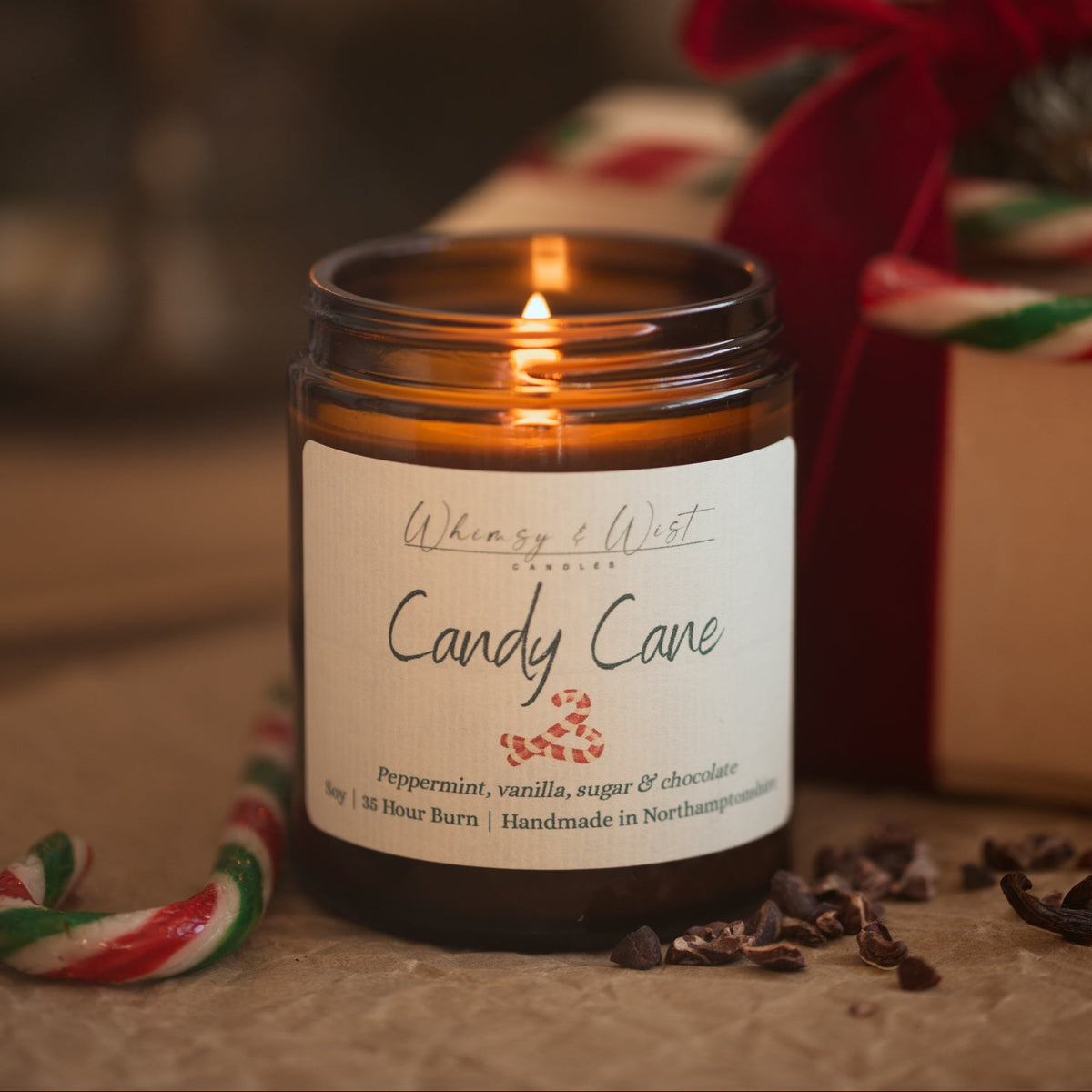 Lit Candy Cane peppermint candle in an amber jar with candy canes, cacao nibs and a gift tied in red ribbon