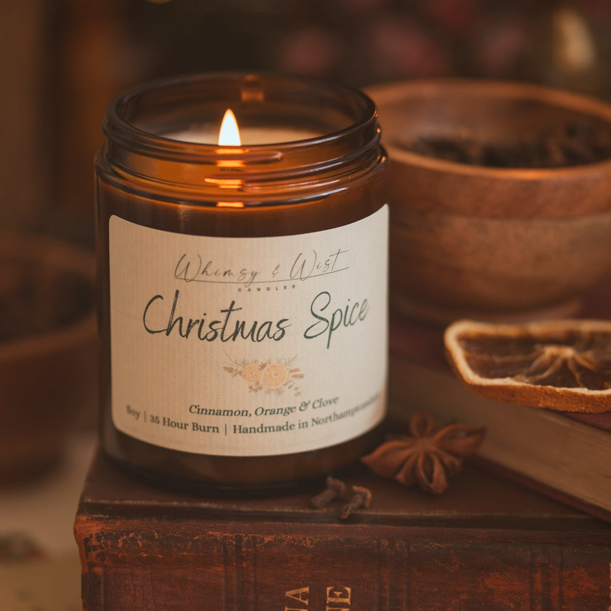 Lit Christmas Spice soy candle on an old book with star anise, dried orange and a wooden bowl