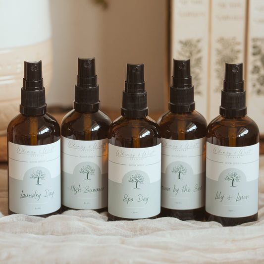 Five amber glass spray bottles with black spray tops and white labels are lined up on a light fabric surface. The background shows a potted green plant and a stack of books.