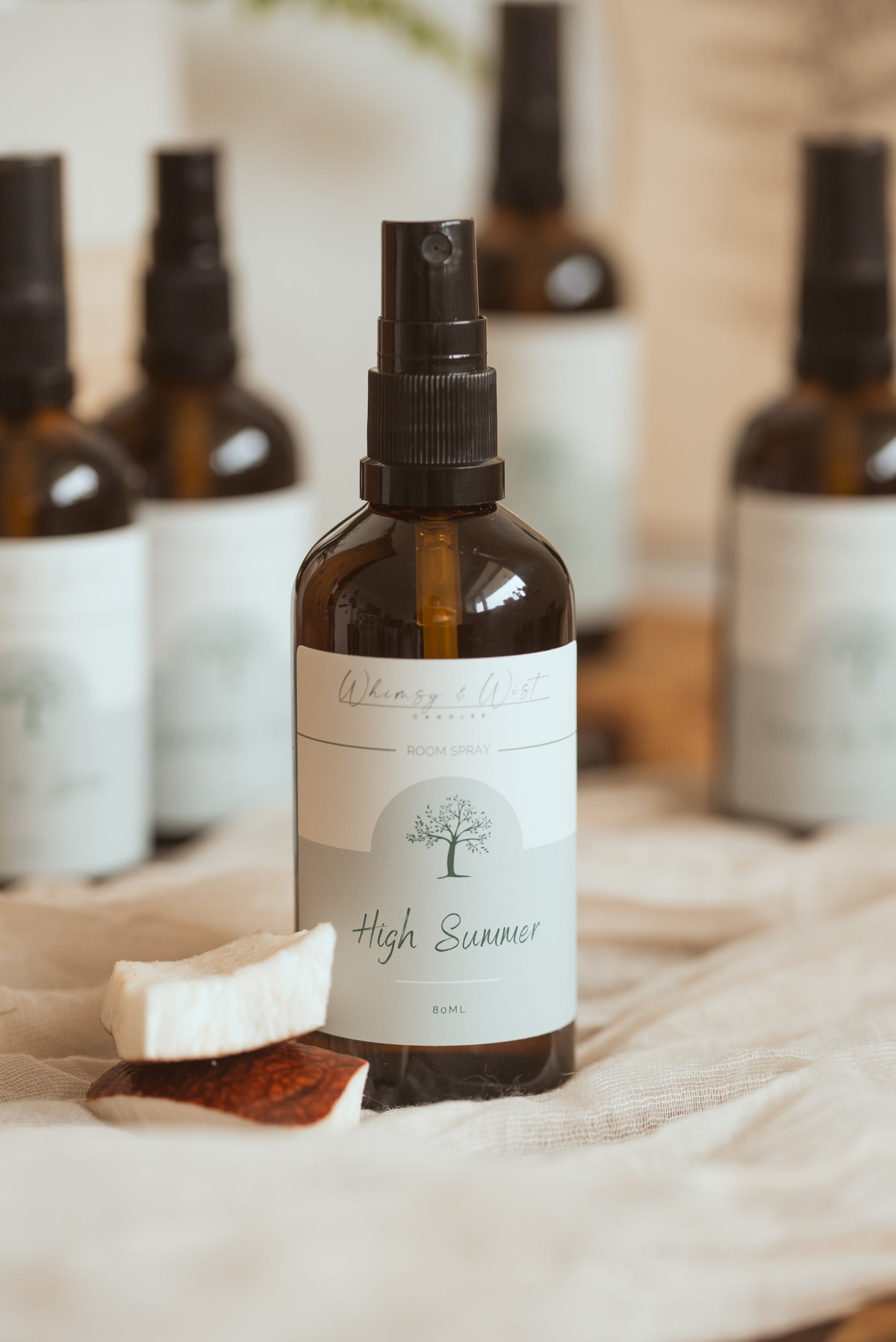 An amber glass spray bottle labeled 'High Summer' by Whimsy & Wist, styled on a beige fabric surface with a soap bar in the foreground and other blurred bottles in the background.