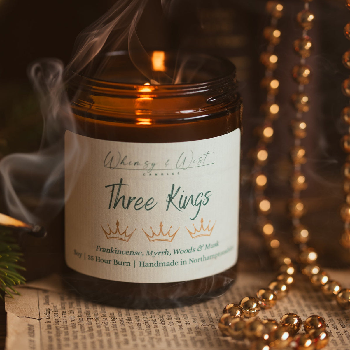 Three Kings soy candle with wisps of smoke curling up beside it, surrounded by gold beads