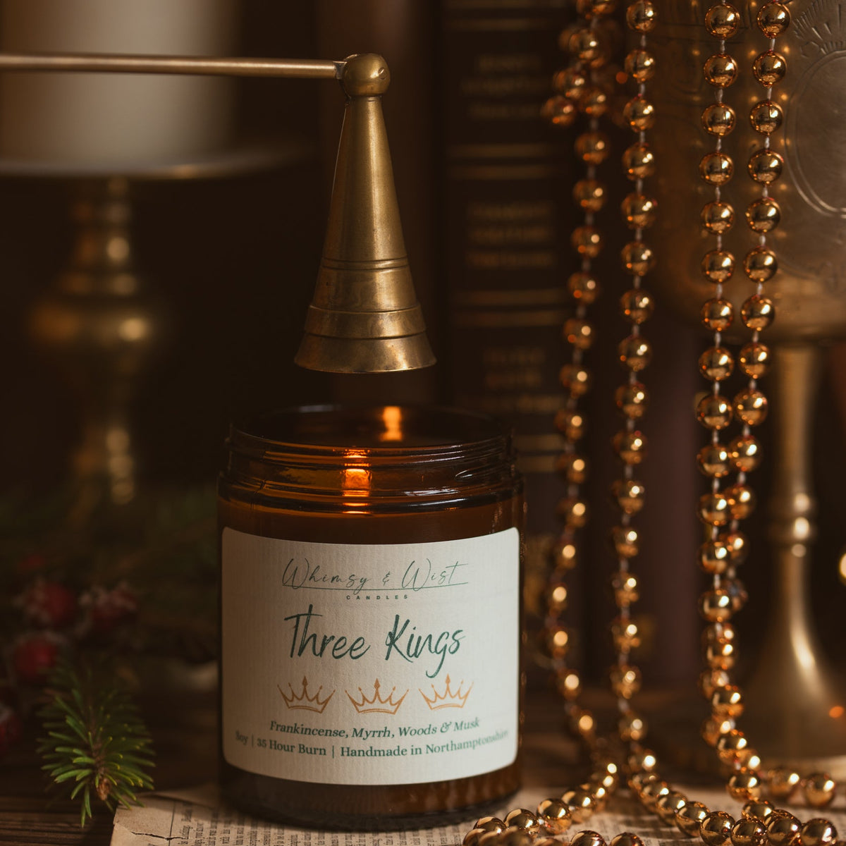 Three Kings soy candle with a brass snuffer above it, gold bead garlands and a brass candlestick behind