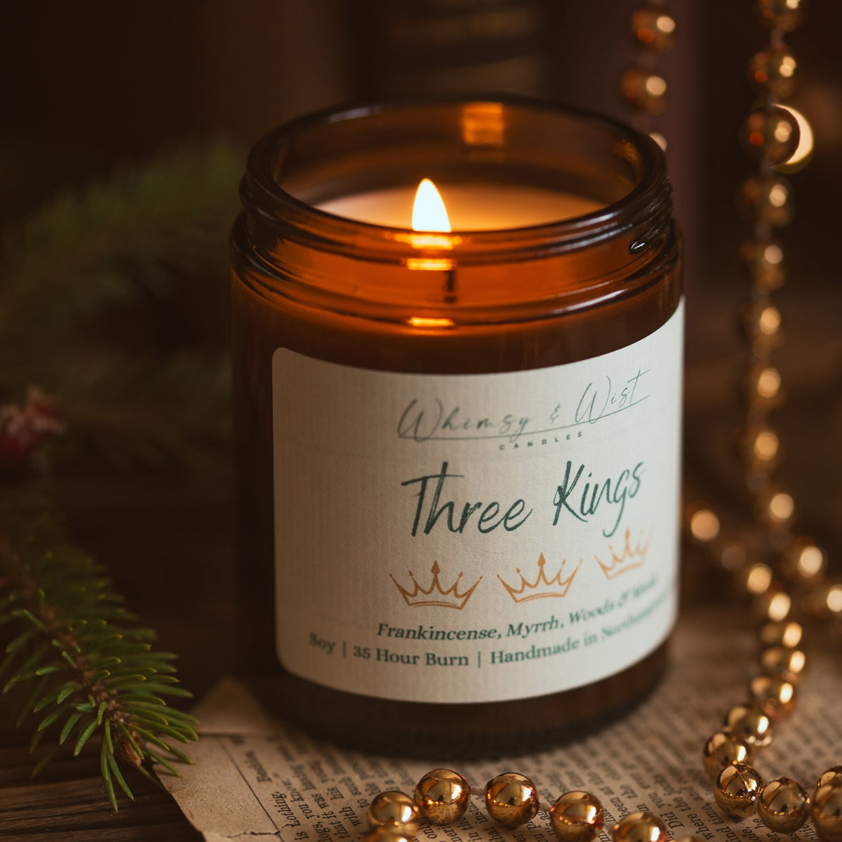 Lit Three Kings soy candle showing the wax pool, with gold beads, pine and vintage book pages