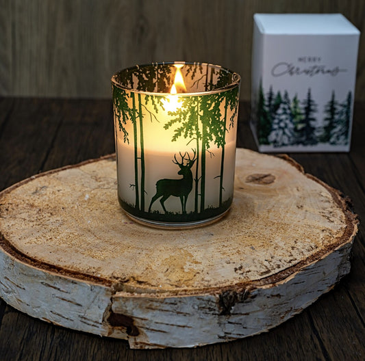 Winter's Tale wood wick Christmas candle lit in a glass jar printed with a green winter forest and stag, beside its Merry Christmas gift box